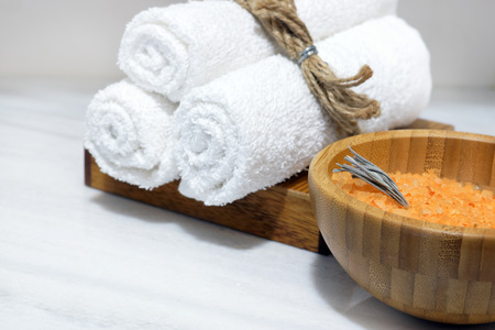 Orange bath salt in a wooden bowl and three white towels on a wooden stand stand on a white marble tableの写真素材