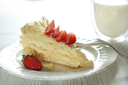 A piece of cake is napoleon made of puff pastry with cream cream, decorated with a flower bed, next to a glass of milk on a table.の写真素材
