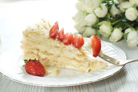 A piece of Napoleon cake on a plate adorned with a red ripe strawberry, a fork with a stuck piece of dessertの写真素材