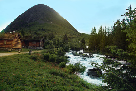 Wooden houses stand near the river against the backdrop of the mountain in Norway. Herbal roofsの写真素材