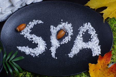 The word spa written in white bath salt on a black dish, next to yellow autumn leaves, palm leaf and acorns. Autumn still lifeの写真素材