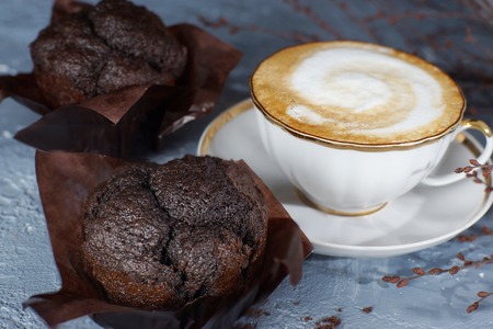 A cup of cappuccino coffee stands on a gray table next to two beautiful chocolate muffins.の写真素材
