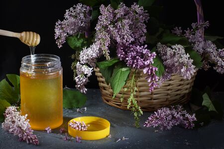 A bouquet of purple lilacs in a wicker basket on a dark background next to a jar of honey and a yellow lid on the tableの写真素材