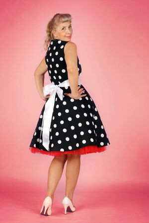 Beautiful blonde woman in a polka-dot dress flirty over her shoulder, pin-up style on a pink backgroundの写真素材