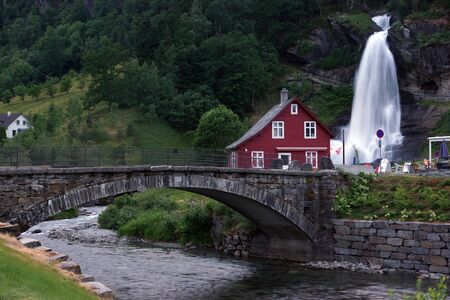 Travel to Norway, a bright house stands near a waterfall, which flows into a river, through which a stone bridge is built.の写真素材