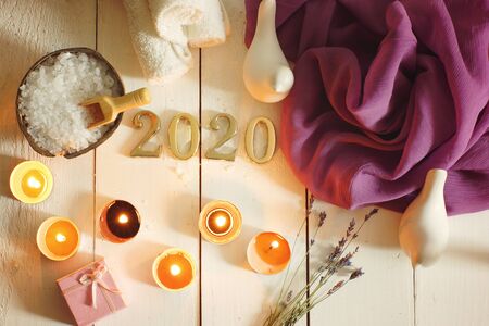 Burning candles illuminate the spa set and gold numbers 2020 on a white plank background with a purple clothの写真素材