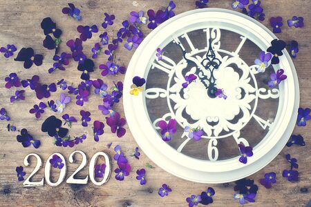 Figures 2020 on a wooden background next to purple pansy flowers and a white clock showing five minutes to midnight.の写真素材
