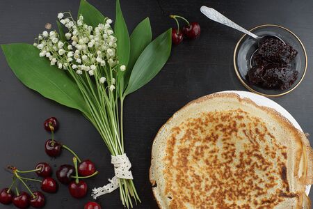 A bouquet of lilies of the valley lies on a table next to pancakes and cherry jamの写真素材