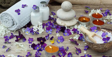 Set for spa treatments with bath salt, stones for hot massage and skin lotions in the midst of small purple flowers of pansies.の写真素材