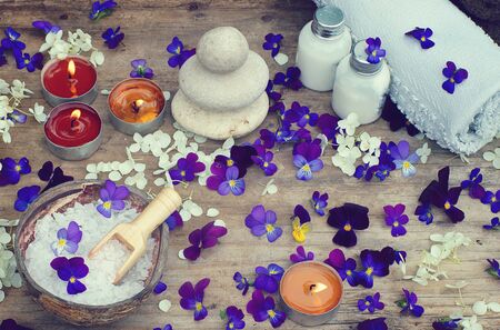 Burning candles and spa set with bath salt, stones for hot massage and skin lotions in the midst of small purple flowers of pansies.の写真素材