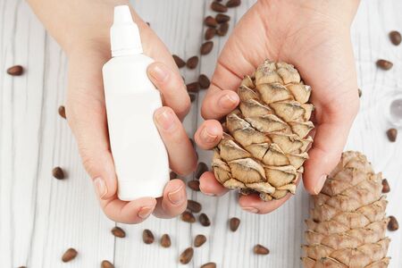 Female hands holding a cosmetic product in a white bottle with an empty label and a pine cone with nuts.の写真素材