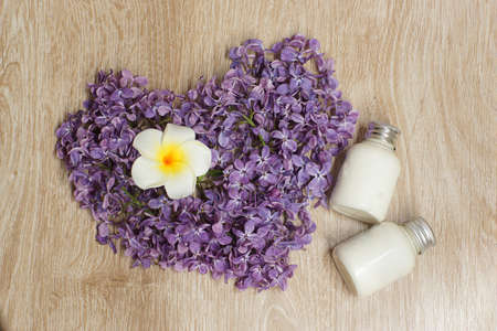 White plumeria flower lies inside a heart made of lilacs on a wooden table, next to cosmetics, top view, spa conceptの写真素材