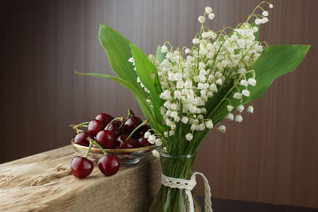 A beautiful bouquet of white lilies of the valley on a brown background is standing next to a bowl with cherries.の写真素材