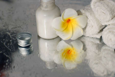 white towel body lotion and plumeria flower are reflected in the mirror surface of the wet table.