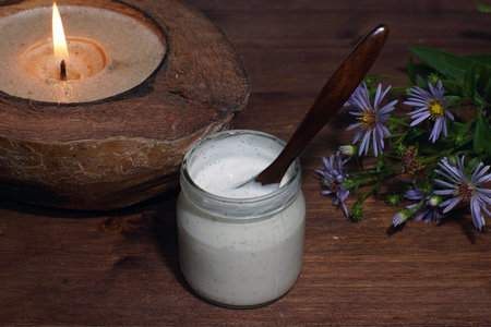 A jar with a scrub or other white cosmetic product is on the table with a burning candle.の写真素材