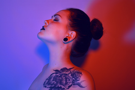 Beautiful young woman with stylish hair and tattoo on her body posing over blue and red background. Cosmetics, make-up. Tattoo. Black lipsの写真素材