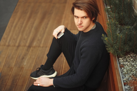 A man sits on a bench with a phone. Fashion man looks into the distance. Fashion man outside in sweater.の写真素材