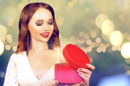 A beautiful brunette opens her New Year's gift against a background of bright lights. The magic of Christmas. A woman in a white dress holds a red box in her hands and smiles at the gift.Professional green makeup and long earrings.の写真素材