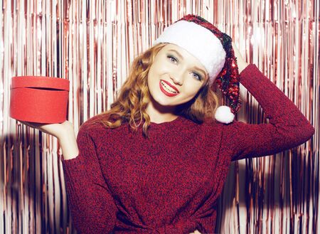 Beautiful girl in a bright santa hat on a shiny background holds a red box with a gift for the new year. Christmas brunette in a red sweater holds a gift and smiles.の写真素材