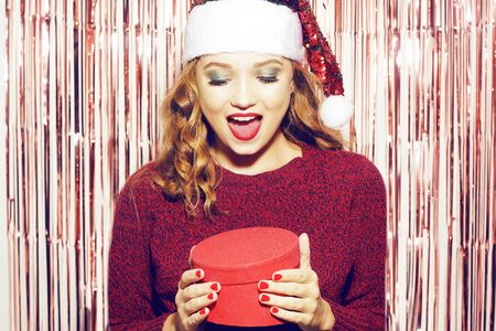 Beautiful girl in a bright santa hat is holding a red box with a gift for the new year. Christmas brunette in a red sweater unpacks her gift and smiles. The girl is looking into the box.の写真素材
