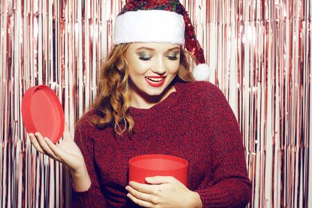 Beautiful girl in a bright santa hat is holding a red box with a gift for the new year. Christmas brunette in a red sweater unpacks her gift and smiles. The girl is looking into the box.の写真素材