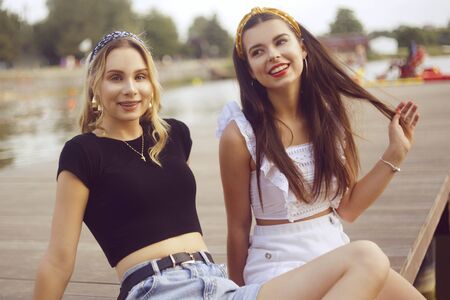 Two fashionable girls, girlfriends, are sitting on the pier near the river, the beach. Girls talk on the beach, laugh, smile, share secrets, discuss guys. Rest in the summer, a trip with a friend.の写真素材