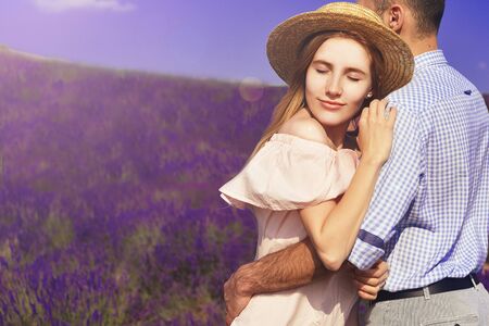 Cute young happy couple in love in a field of lavender flowers. Enjoy a moment of happiness and love in a lavender field. Boy smile to his girl. Blond girl in hat and pink dress. Romantic couple.の写真素材