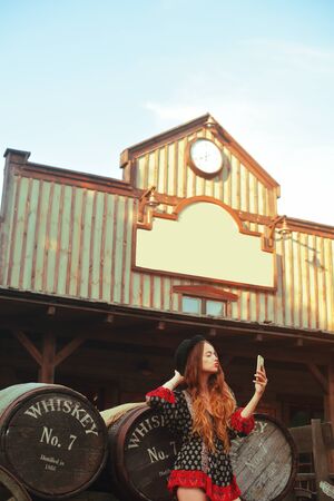 Girl in the Wild West, in Western house. Girl in hat with long cerly hair. Beautiful pretty girl looks into the phone. Incredible trip, travel. Cute woman in red dress pouse.の写真素材