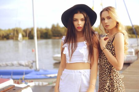 Two girls on the background of yachts, sailing boats are smiling, looking at the camera. Brunette in a hat and blonde in a dress with a leopard print on vacation. Travel, exchange students.の写真素材