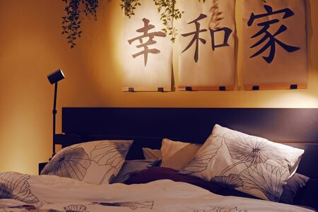 Japanese interior. Painting with hieroglyphs. Bed and pictures above it. Evening room background. Bedroom, pillows on the bed.の写真素材
