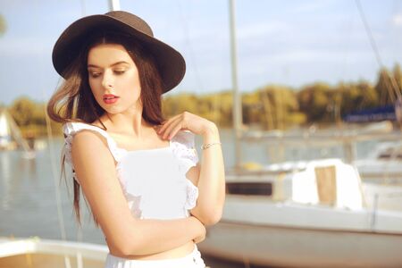 Girls on the background of yachts, sailing boats smiling, looking at the camera. Beautiful brunette in a hat and jacket on vacation. Fashionable girl in summer. Relaxation.の写真素材