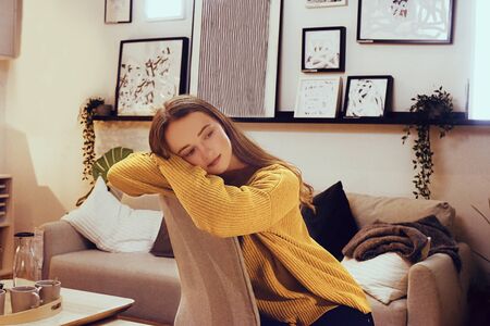 A sad girl is sitting in a room on an armchair, sad. girl in a bright yellow sweater on the background of photo frames and sofaの写真素材