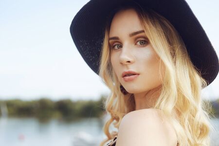 Girl on background of yachts, sailing boats smiling, looking at the camera. beautiful blonde is sitting on the pier, in a dress with a leopard print and a hat on vacation. Fashionable girl in summerの写真素材