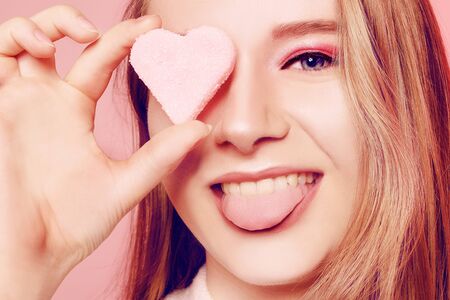 Valentine's Day, a girl on a pink background, closes her eye with a heart. The blonde smiles beautifully, laughs, shows her tongue, makes faces, has fun, love. March 8, gifts for the girl.の写真素材