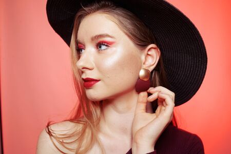 Gorgeous blonde in a big hat with burgundy make-up, red lips, and a burgundy dress. Girl in burgundy earrings, portrait of a model. Fashion photo of bright red makeup, model on a red backgroundの写真素材