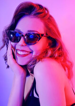 Beautiful girl in neon. Fashion photo of a short-haired girl, dancing, sun glasses. Girl in red and blue light posing. Happy girl smiles, laughs, good emotions.の写真素材