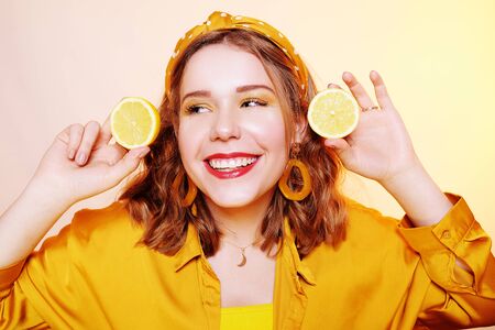 Beautiful girl, yellow make-up, bright colors. Girl holds lemons near her face, smiling. Facial masks made of lemon, procedures with lemon. Girl with accessories, big earrings, headband.の写真素材