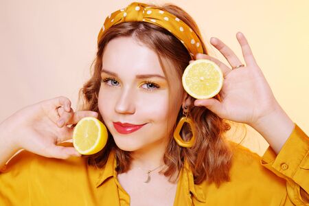 Portrait of a girl, yellow makeup, bright colors. Girl holds lemons near her face, smiling. Girl with accessories, big earrings, headband, yellow background.の写真素材