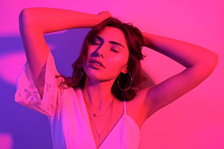 Brunette with long curled hair dancing in neon. A girl with professional makeup in red and blue light posing. Sexy model in a white dress at a party.の写真素材