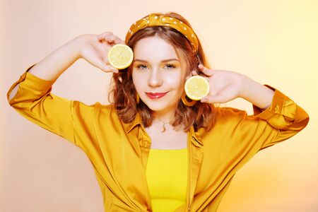 Beautiful girl, yellow make-up, bright colors. Girl holds lemons near her face, smiling. Facial masks made of lemon, procedures with lemon. Girl with accessories, big earrings, headband.の写真素材