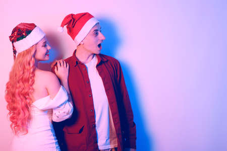 Couple in santa hats, new year, christmas. Lovers in neon light. Surprised couple looking to the side at empty space for textの写真素材