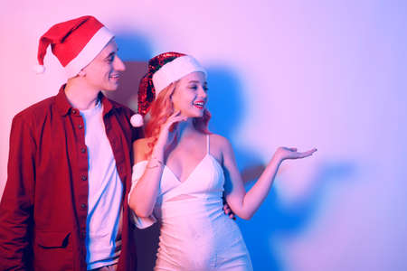Couple in santa hats, new year, christmas. Lovers in neon light. Surprised couple looking to the side at empty space for textの写真素材