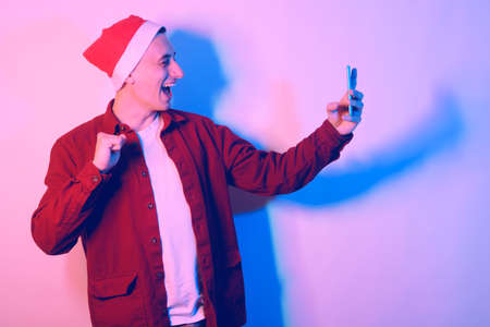 A man in a santa hat in neon light. The guy is talking by video with his family. New Year's and...