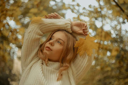 Photo portrait of a girl in a white sweater, the blonde holds leaves in her hands. Pretty woman in the park in autumn. Autumn fashion and style. Warm moodの写真素材