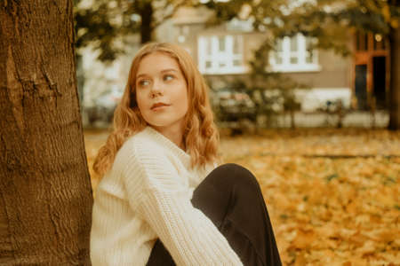 A girl in a white sweater sits on the autumn leaves. A beautiful blonde with a square looks at the...