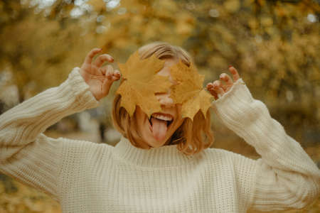 Photo portrait of a girl in a white sweater, the blonde holds the leaves and closes her eyes. Pretty woman in the park in the fall shows her tongue, joy, smile. Autumn fashion and styleの写真素材