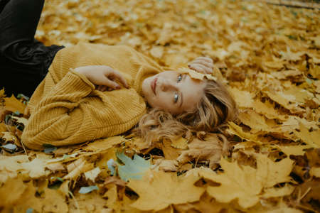 Beautiful girl lies in the park in autumn. A teenager in a yellow sweater on a background of orange leaves. The blonde looks at the camera. Fashion and Styleの写真素材