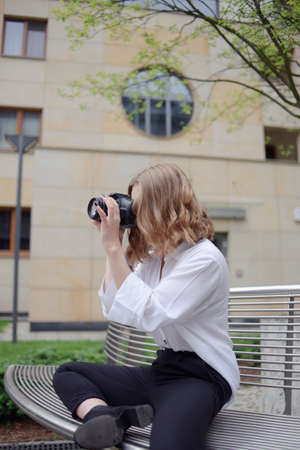 A beautiful girl looks in the viewfinder in the camera, takes pictures. Blonde learns to photograph and earn money on the photo. Photographer walks around city, takes pictures of everything around.の写真素材