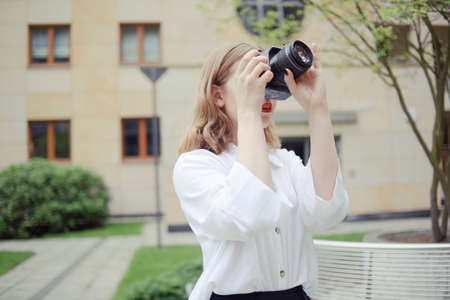 A beautiful girl looks in the viewfinder in the camera, takes pictures. Blonde learns to photograph and earn money on the photo. Photographer walks around city, takes pictures of everything around.の写真素材