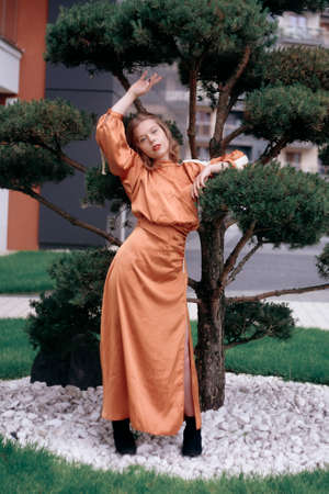A girl in a long evening brown dress stands near a tree. Professional model posing near a tree, blonde with red lips in a fashionable tint.の写真素材